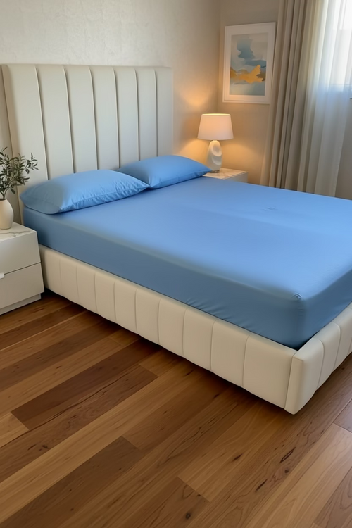 A queen sized bed in a light blue sheet and two matching blue pillows, set in a modern bedroom with a cream tufted platform bed frame, a lit bedside lamp, and wooden floor