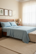 A neatly made bed in a softly lit bedroom, dressed with light blue striped bedding and matching pillows, with a beige rug, cream curtains, and a tufted bench at the foot of the bed.
