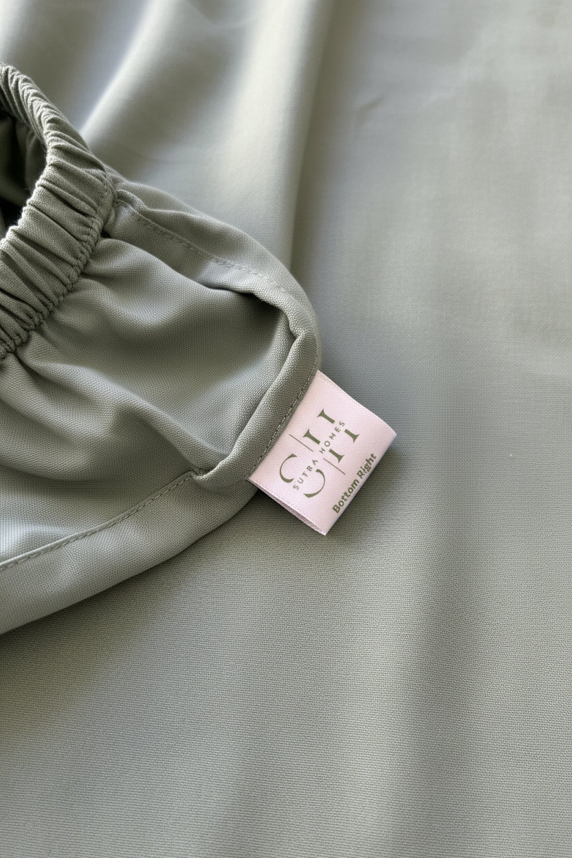 Close-up of a sage green fitted sheet with an elasticized waistband and a sewn-in white label reading 'Bottom Right' for ease