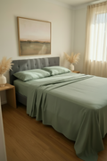 A neatly made bed in a softly lit bedroom, dressed with sage green bedding and matching pillows, with a gray upholstered headboard, wood floor, side tables, and a framed landscape painting above.