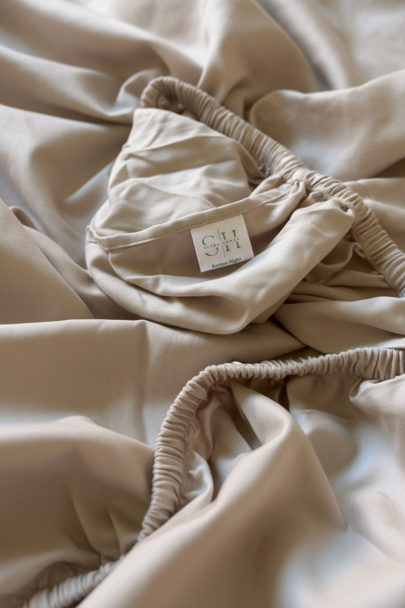 Close-up of a beige or oatmeal-colored fitted sheet with elasticized ruching and a small sewn label reading 'SH' & 'Bottom Right' on a soft, draped background.