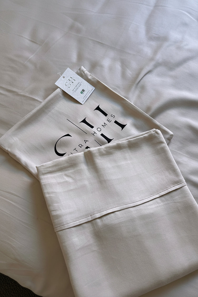 A folded beige cotton bedding set, including a pillowcase and a matching pillow, displayed on a matching bed sheet. The pillowcase features a black printed logo for 'Sultra Homes'.