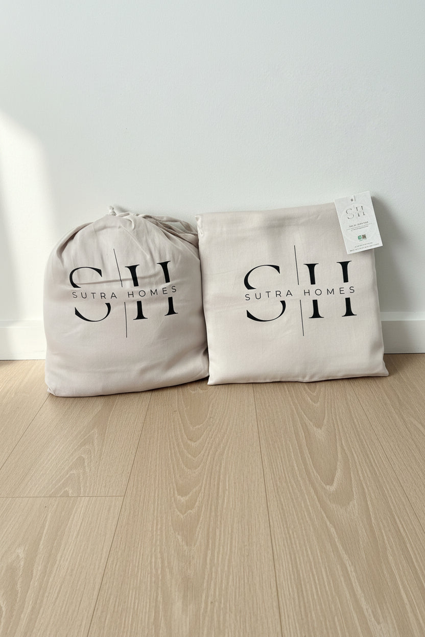 Two light beige fabric storage bag containing either flat or fitted sheet and 2 pillow caseswith black printed 'SUTRA HOMES' logos, one drawstring-top bag and one flat bag (envelope closure) with a hangtag, placed on a against a white wall.