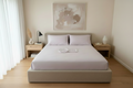 A modern bed with a low upholstered frame, dressed in light lavender or a muted lilac bedding and two matching pillows, set in a minimalist bedroom with light wood flooring, white curtains, and neutral-toned wall art.