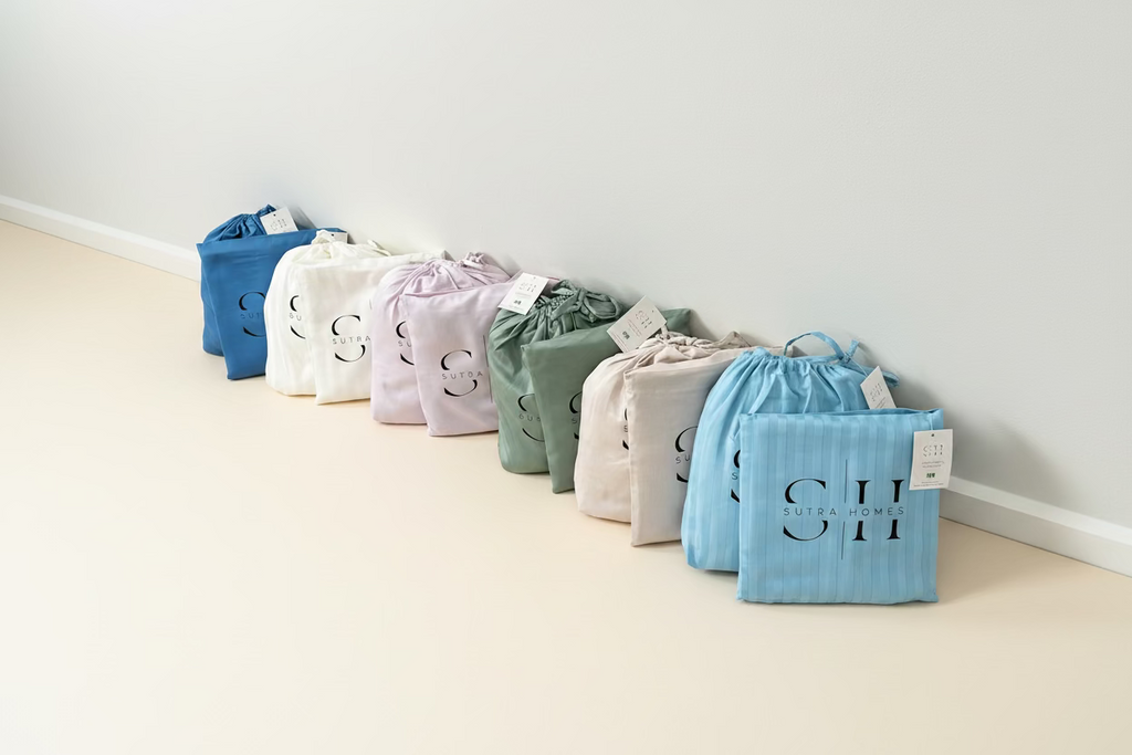 A row of all 300 TC long-staple cotton sateen sheets with pillows in a drawstring storage bags in multiple colors lined up on against a white wall. The sheets are in shades of blue, white, lavender, sage green, cream, and light blue, each printed with a large black monogram 'SH' highlighting the brand Sutra Homes.
